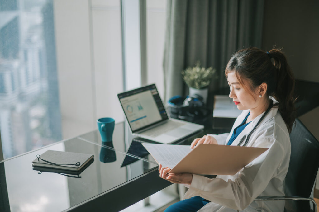 Asian female doctor working on medical report with laptop and document file