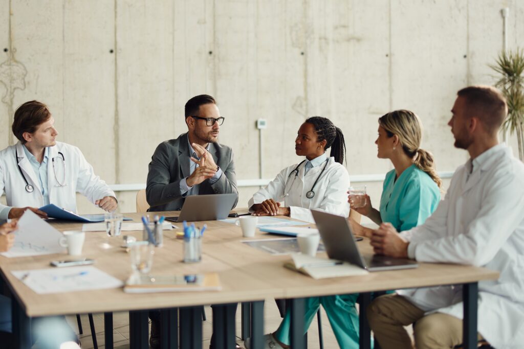 Doctors and a business professional sitting around a table in discussion, with laptops, documents, and coffee cups in front of them.