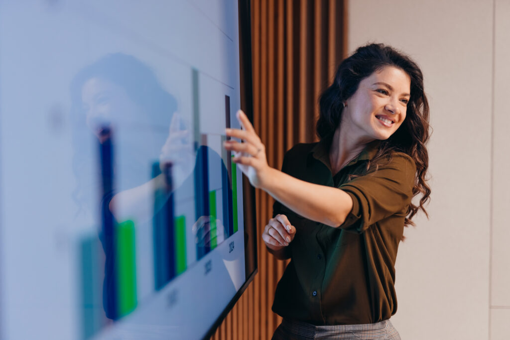 Young businesswoman pointing at an interactive whiteboard during an engaging office presentation, showcasing positive results while smiling confidently and inspiring her colleagues